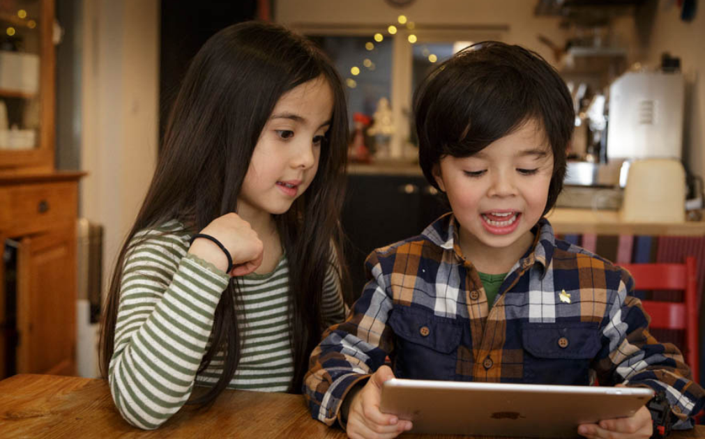 A photo of a brother and sister sitting at their kitchen table. The boy is talking into an iPad, while his sister looks over his shoulder.