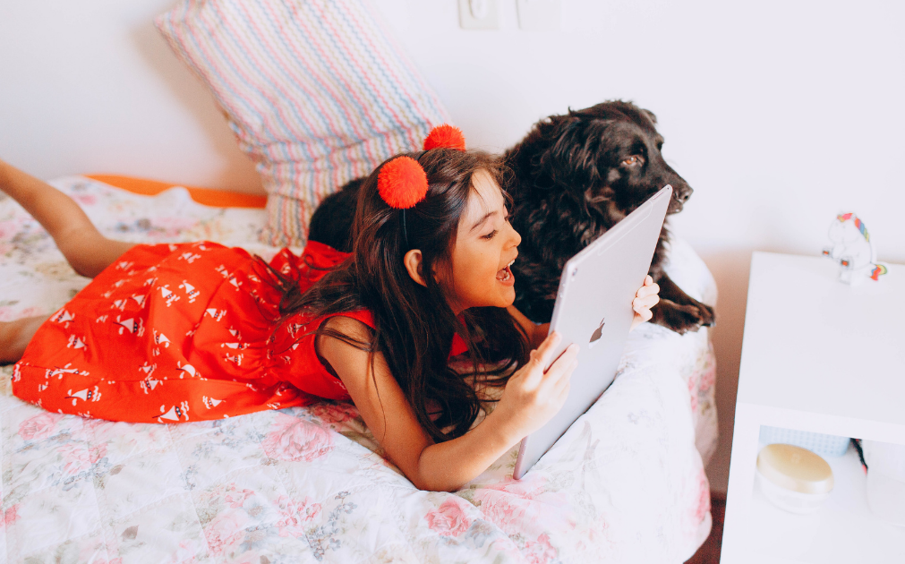 A photo of a young girl lying on her bed, talking into an iPad. A dog is lying next to her.