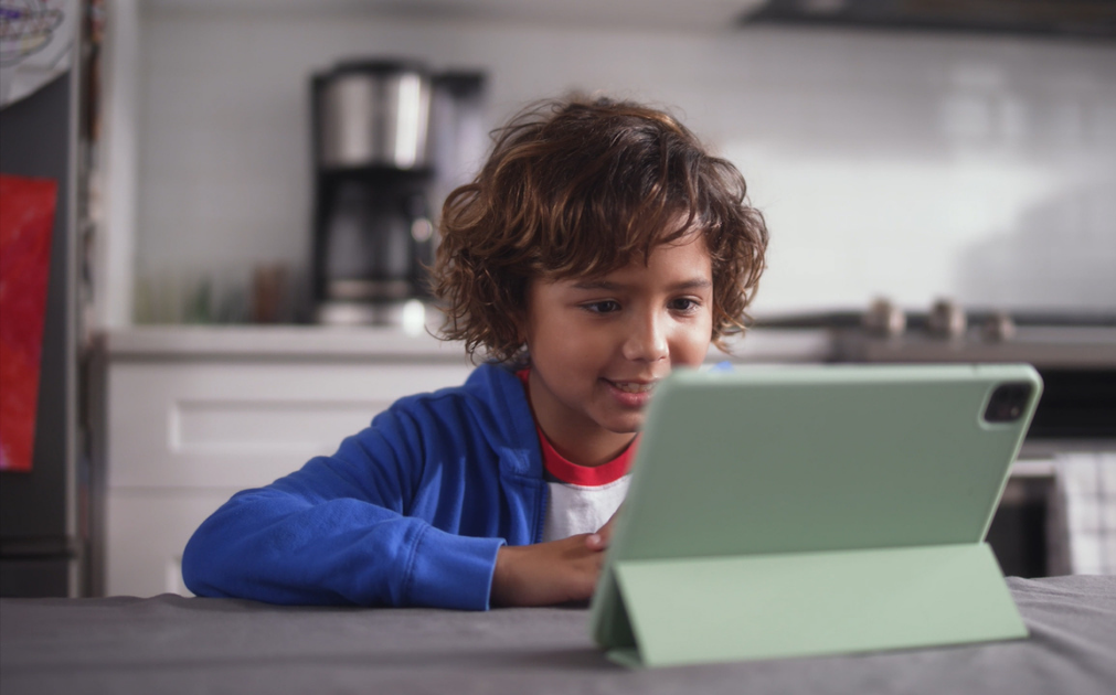 A photo of a boy sitting at a kitchen table talking into an iPad.