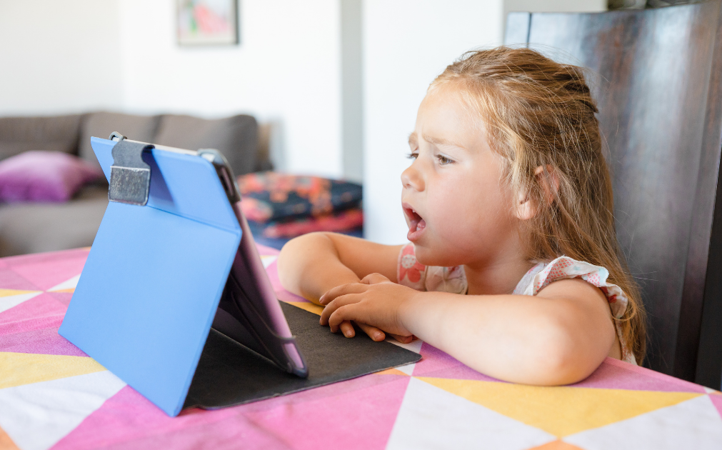 A photo of a young girl setting at a kitchen counter, speaking into a tablet.