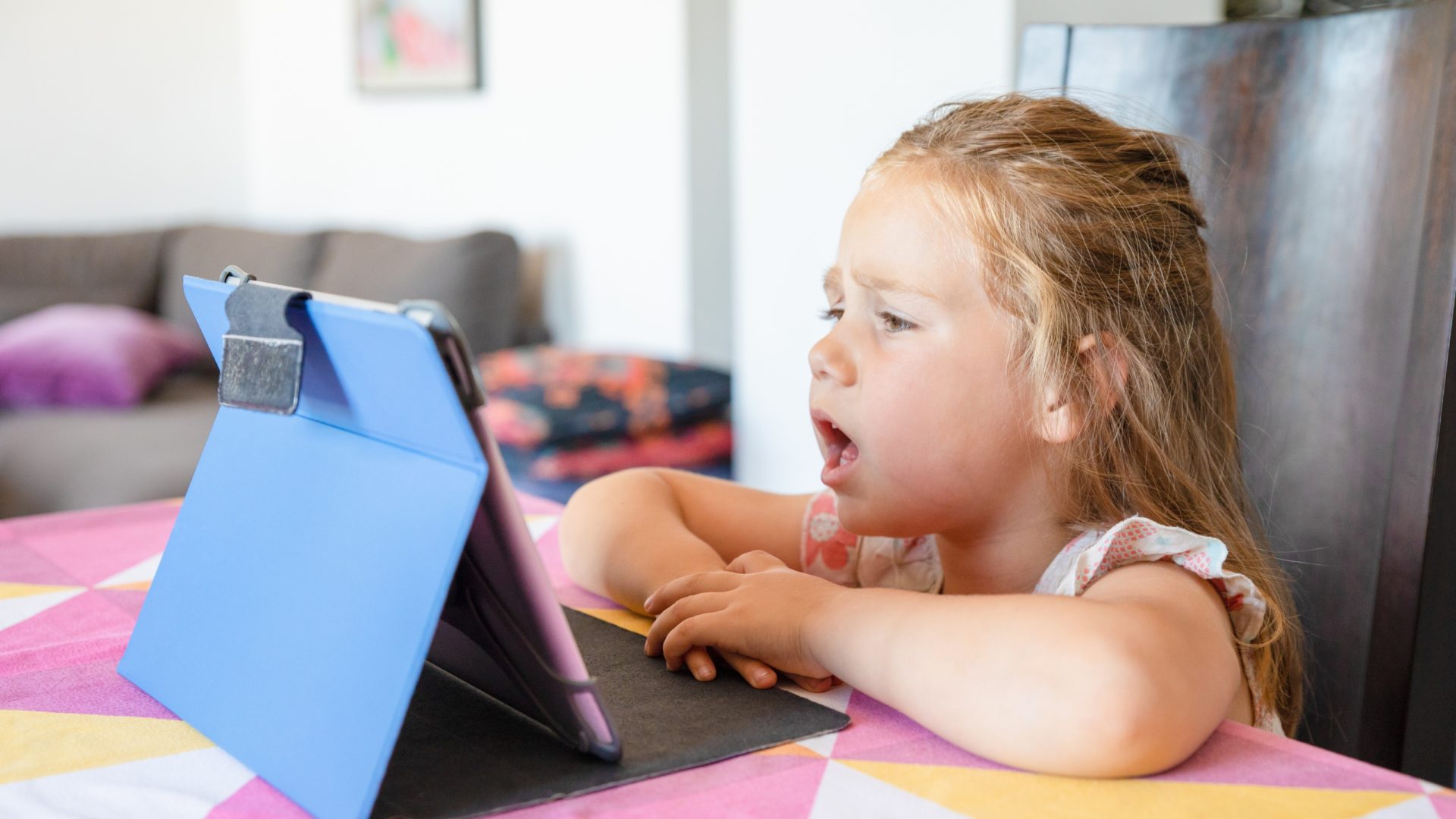 A photo of a young girl setting at a kitchen counter, speaking into a tablet.