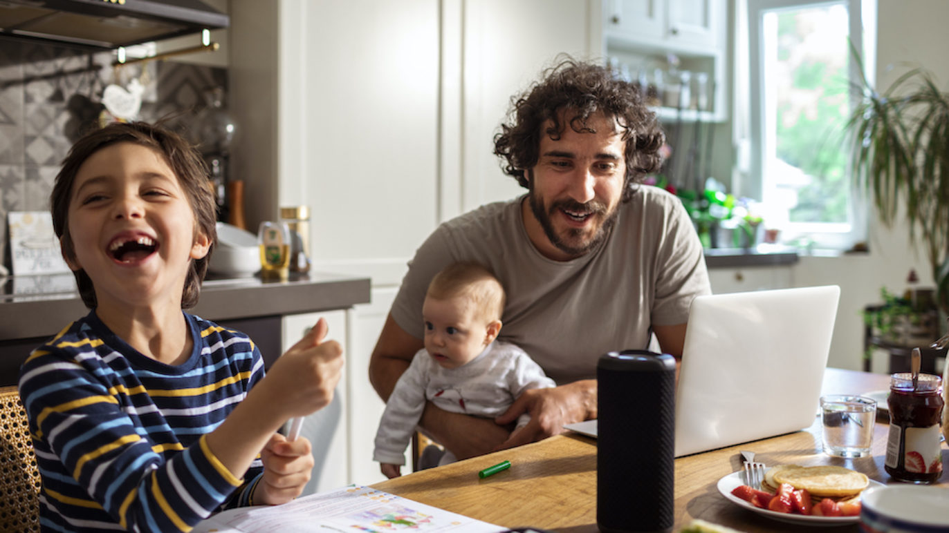 A photo of a dad, his young son, and baby sitting at a kitchen table talking to a smart speaker.