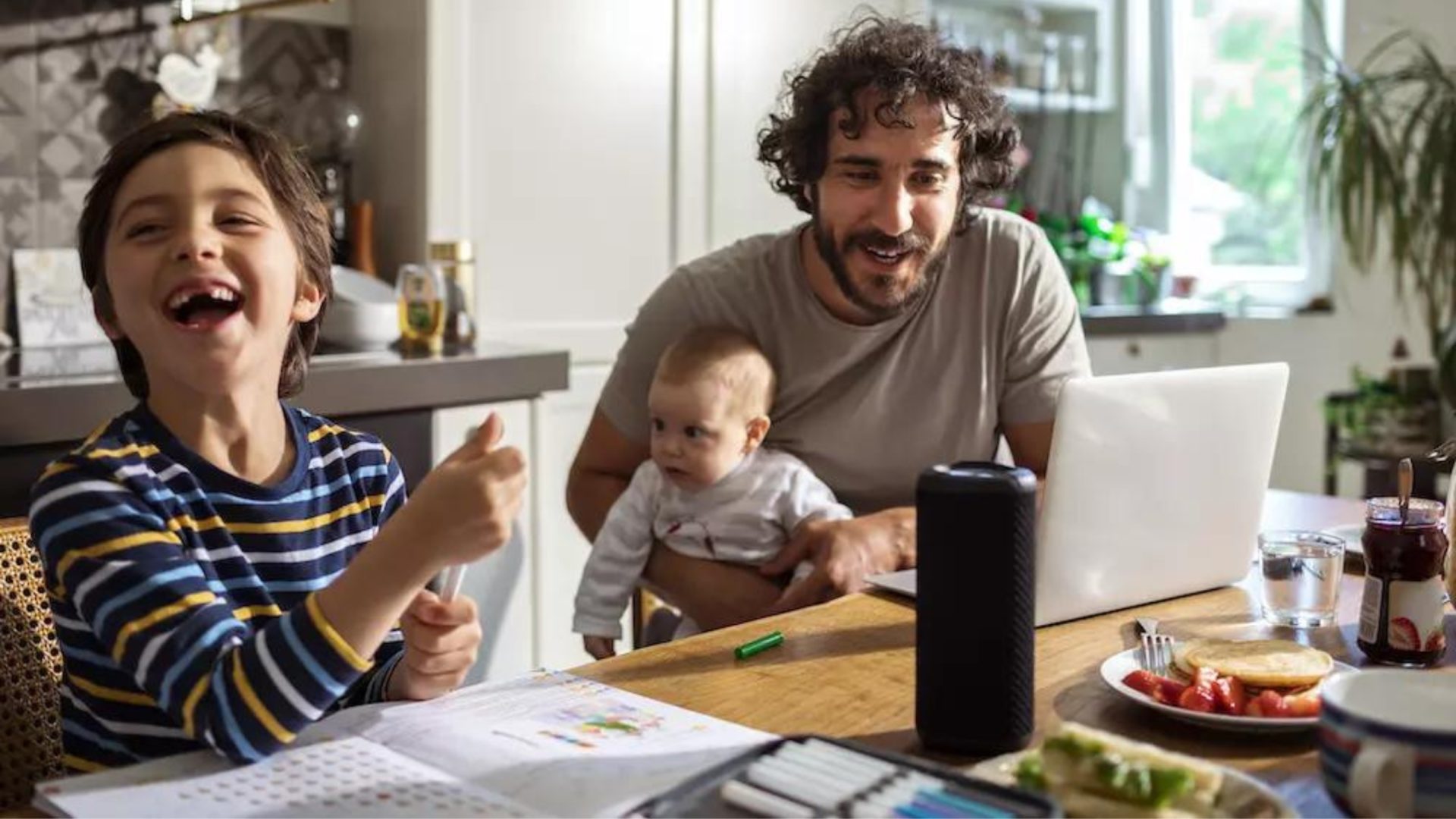A photo of a dad, his young son, and baby sitting at a kitchen table talking to a smart speaker.