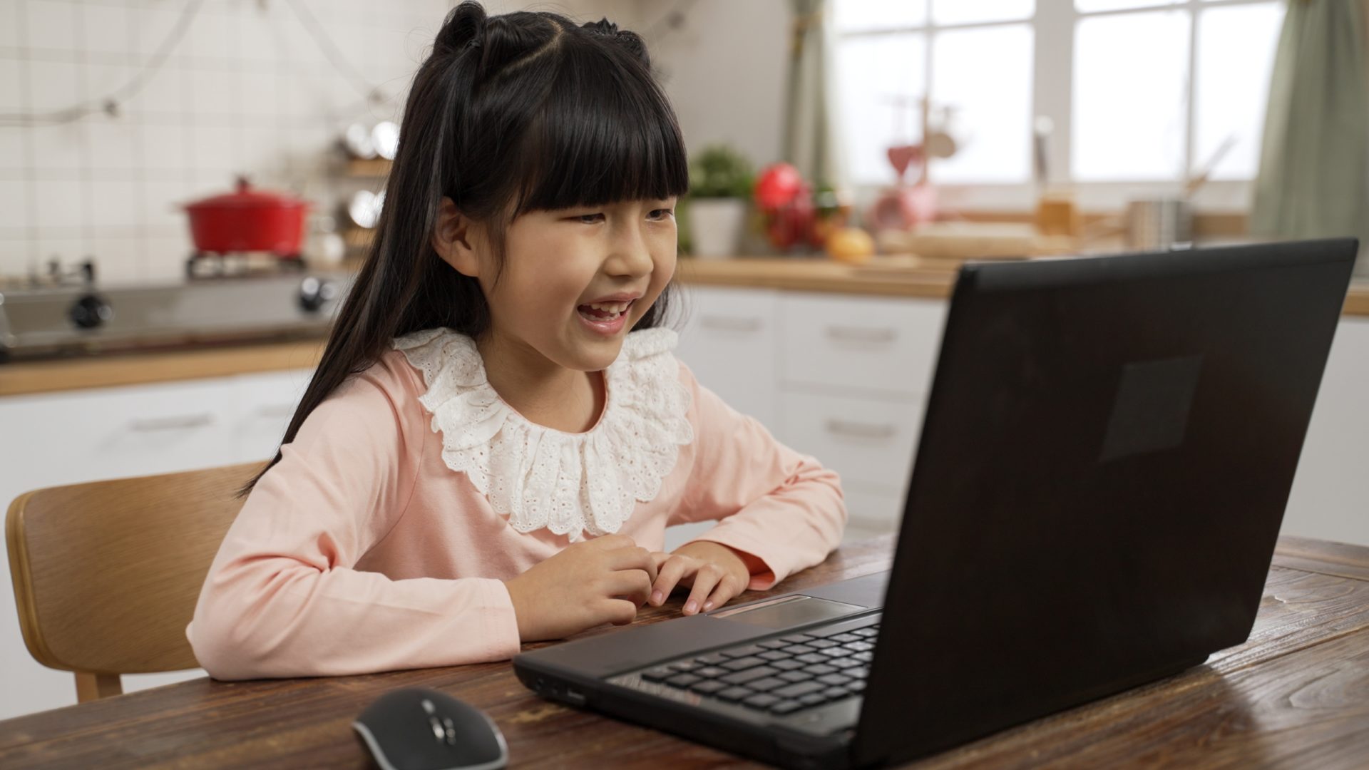 A photo of a girl sitting at a kitchen table, completing a voice-enabled literacy assessment on a laptop computer.