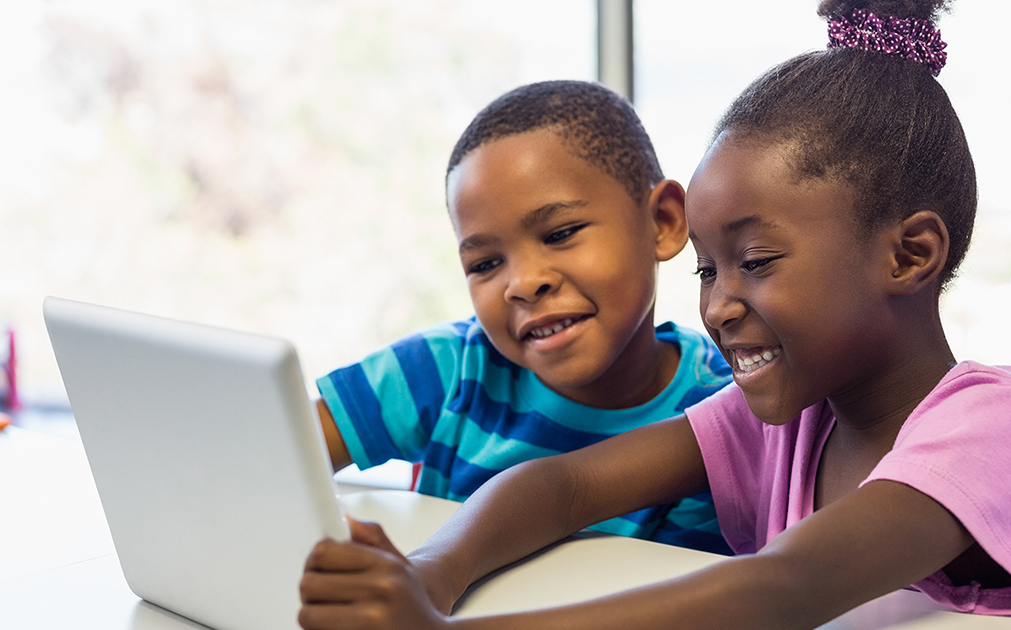 A photo of two young kids, a boy and a girl, sitting at a table reading off of a tablet.