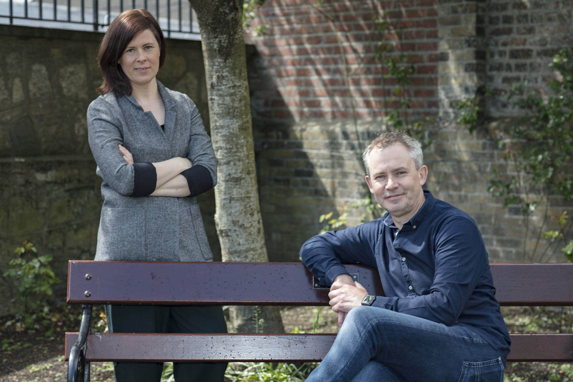 Photo of a man and a woman. Man is sitting on a bench, and woman is standing behind the bench.