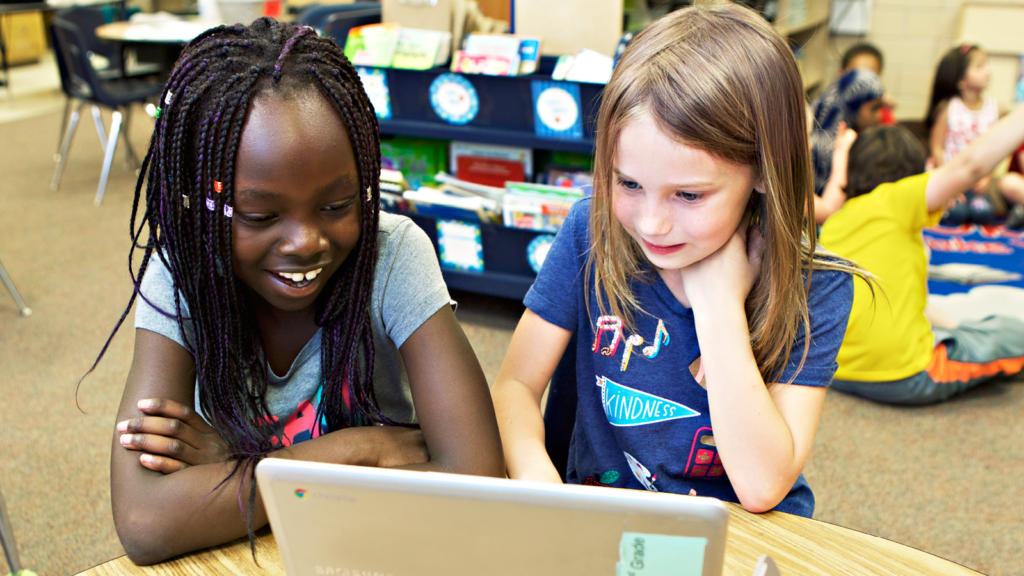 A photo os two girls sitting at a desk in a classroom, reading off of an ipad.
