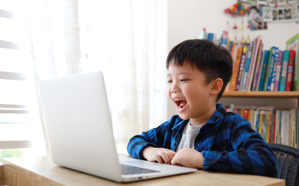 A photo of a young boy sitting at his desk speaking into his laptop.