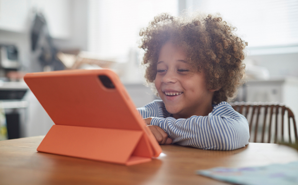 A photo of a young boy sitting at a kitchen table, smiling while looking at his iPad.