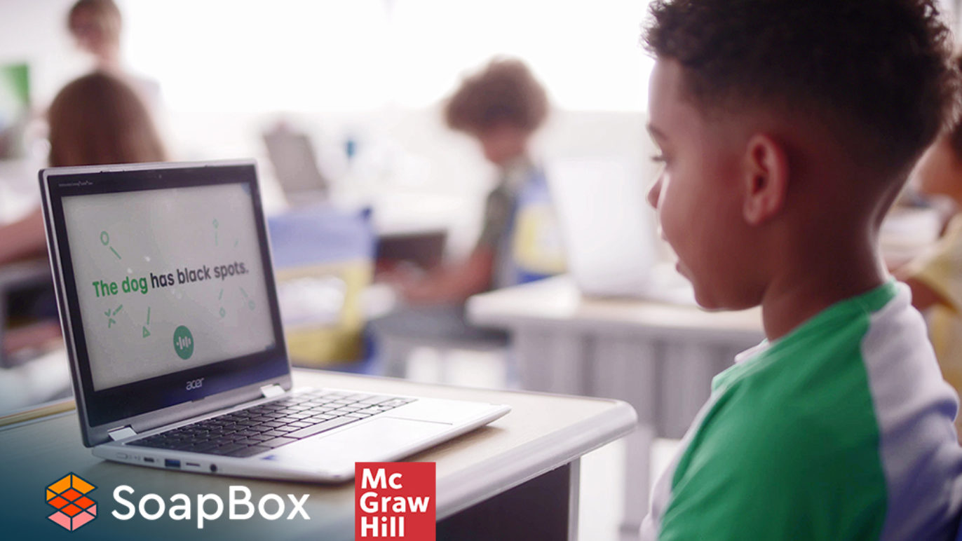 A photo of a young boy sitting at a desk in a classroom, talking into a computer.