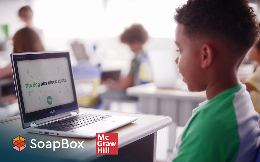 A photo of a young boy sitting at a desk in a classroom, talking into a computer.