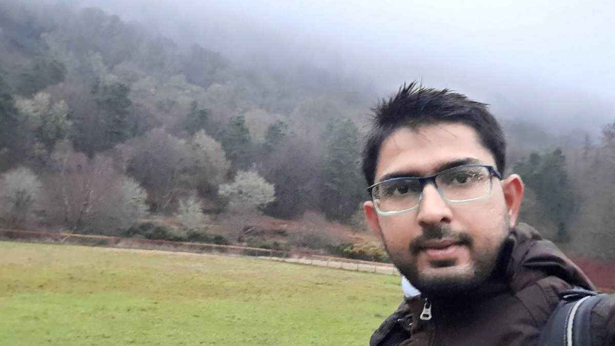 A photo of a man standing outside, with a green field and trees behind him.