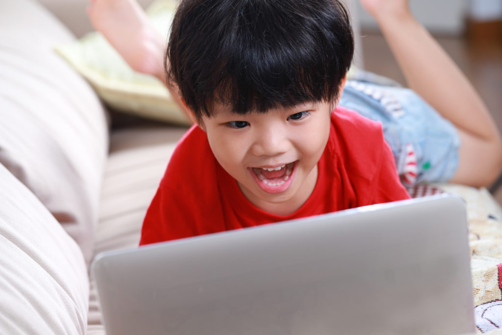 A boy lying on a couch, talking into a laptop computer.