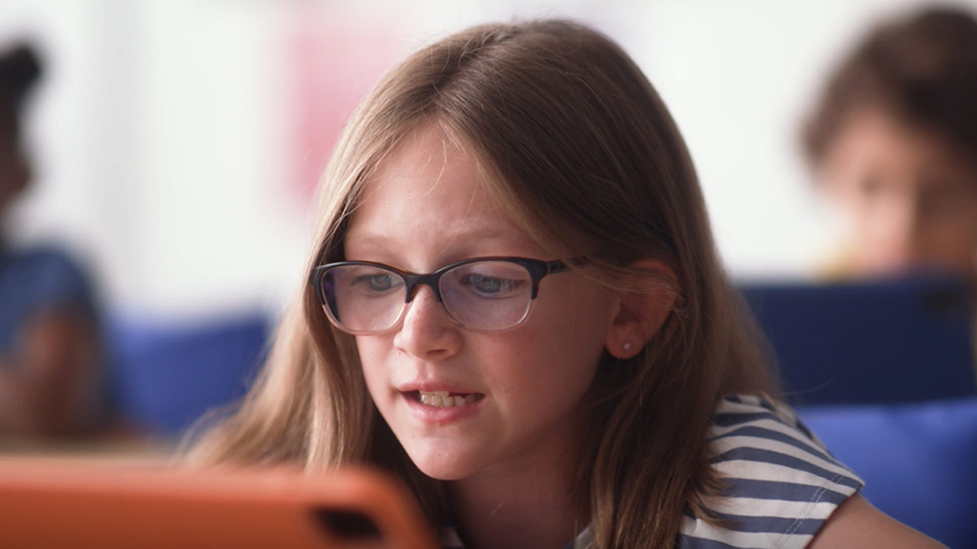 An image of a young girl sitting in a classroom, talking into an iPad.