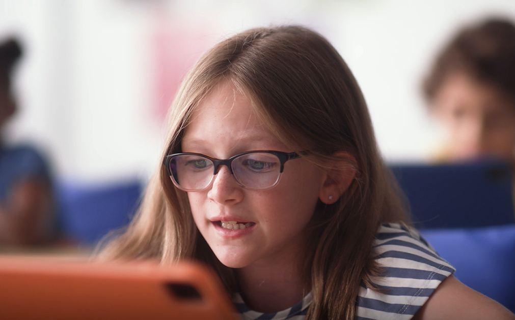 An image of a young girl sitting in a classroom, talking into an iPad.