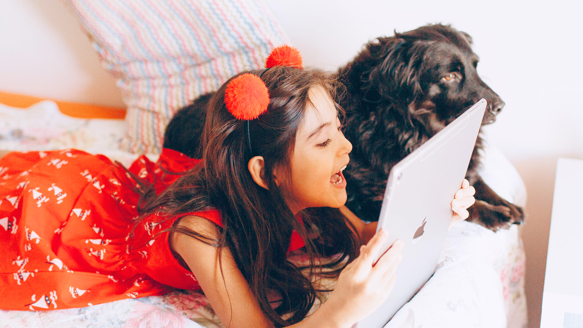 A girl lying on a bed, speaking into an iPad. A dog is lying beside her. 