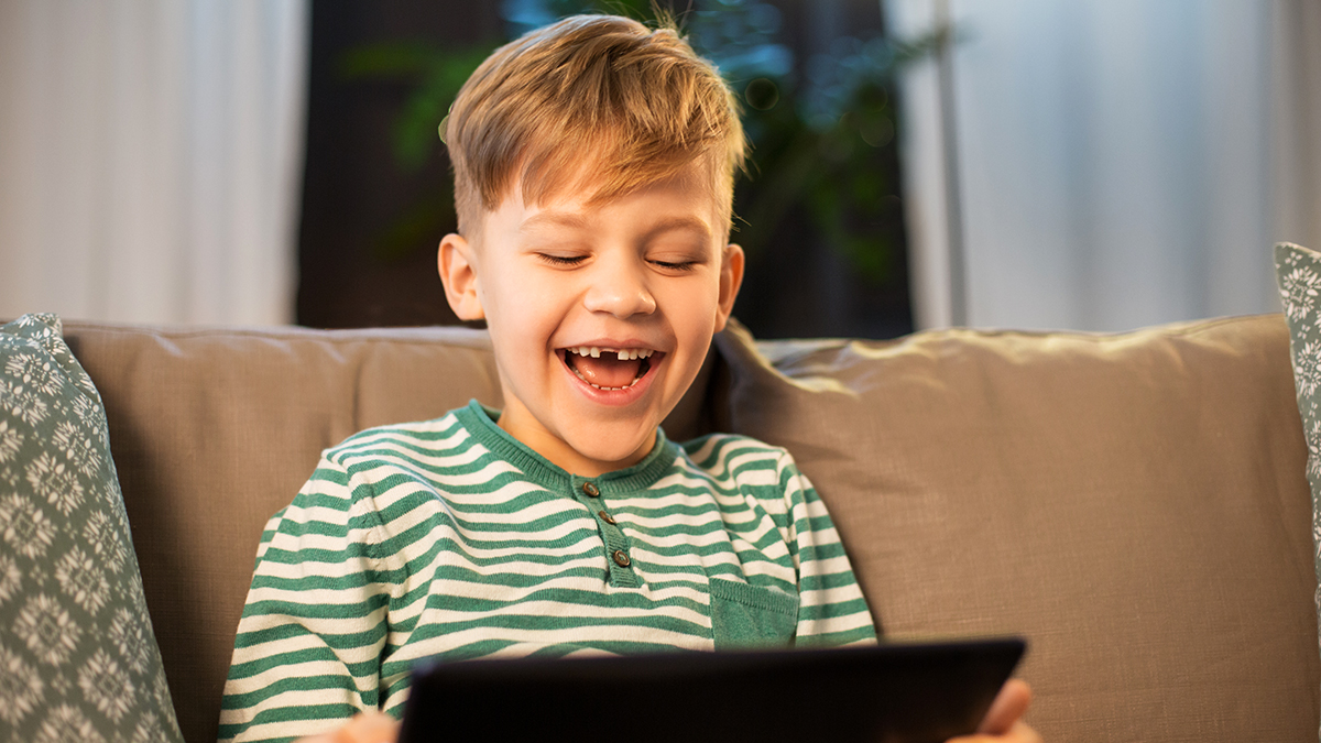 A boy smiling while sitting on a couch, talking into a tablet computer.