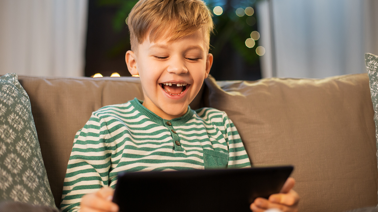 A boy with a big smile sitting on a couch playing on a tablet computer.
