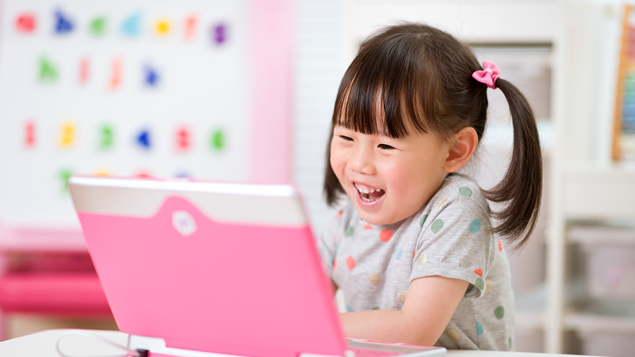 A girl sitting at a desk, smiling and talking into her laptop computer.