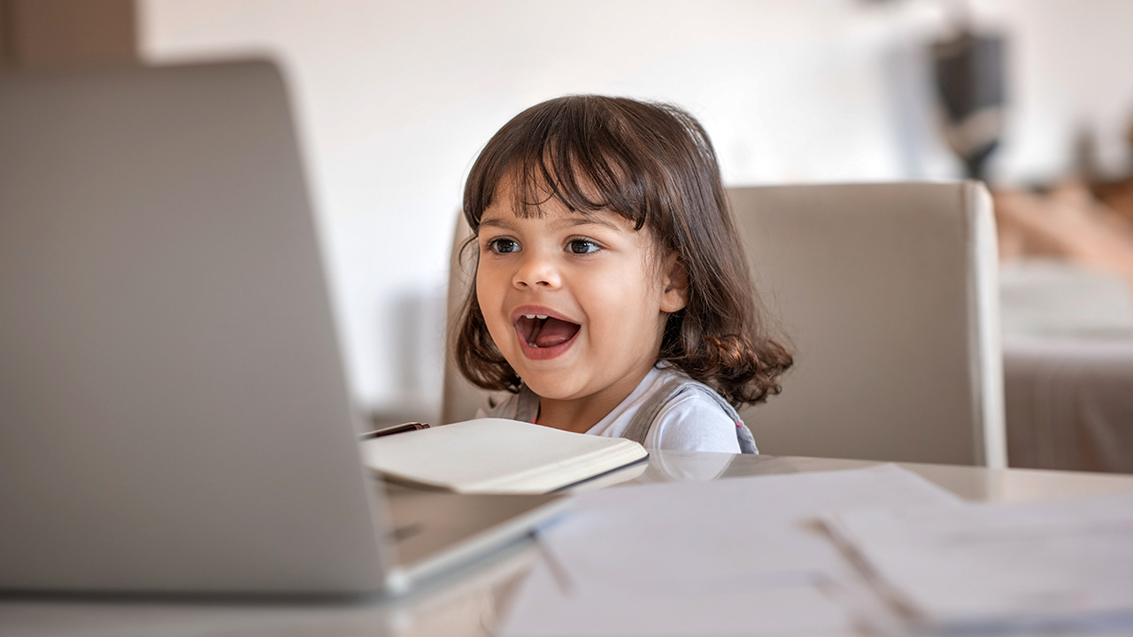 A young girl sitting at a table, talking into a laptop computer.