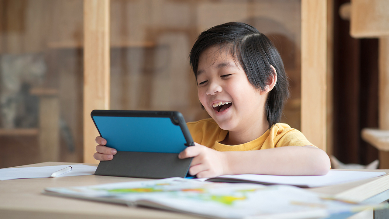 A boy laughing while playing with his tablet computer at a table.