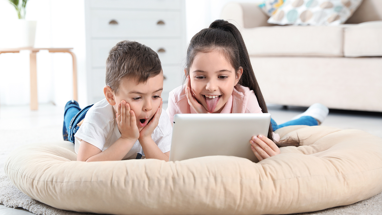 Two kids, a boy and a girl, lying on a big cushion and laughing at what they see on a tablet.