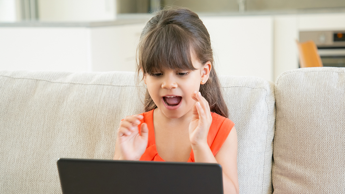 A photo of a young girl sitting on a couch and talking into a laptop computer.