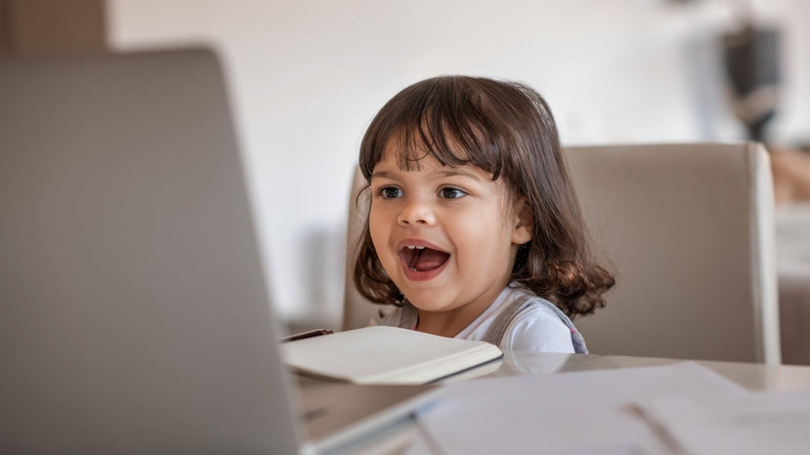 A photo of a girl sitting at a table, talking into a laptop computer and practicing her early literacy skills.