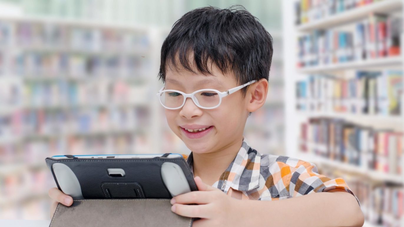 A photo of a boy sitting in a library practicing his oral reading fluency with a voice-enabled reading app.