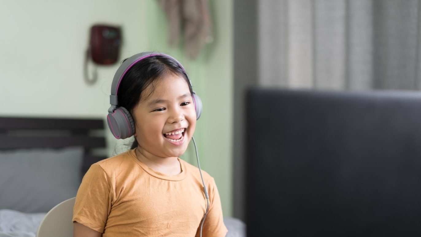 An photo of a girl sitting at a desk in her bedroom. A laptop computer is in front of her, and she is wearing big headphones. The girl is smiling.