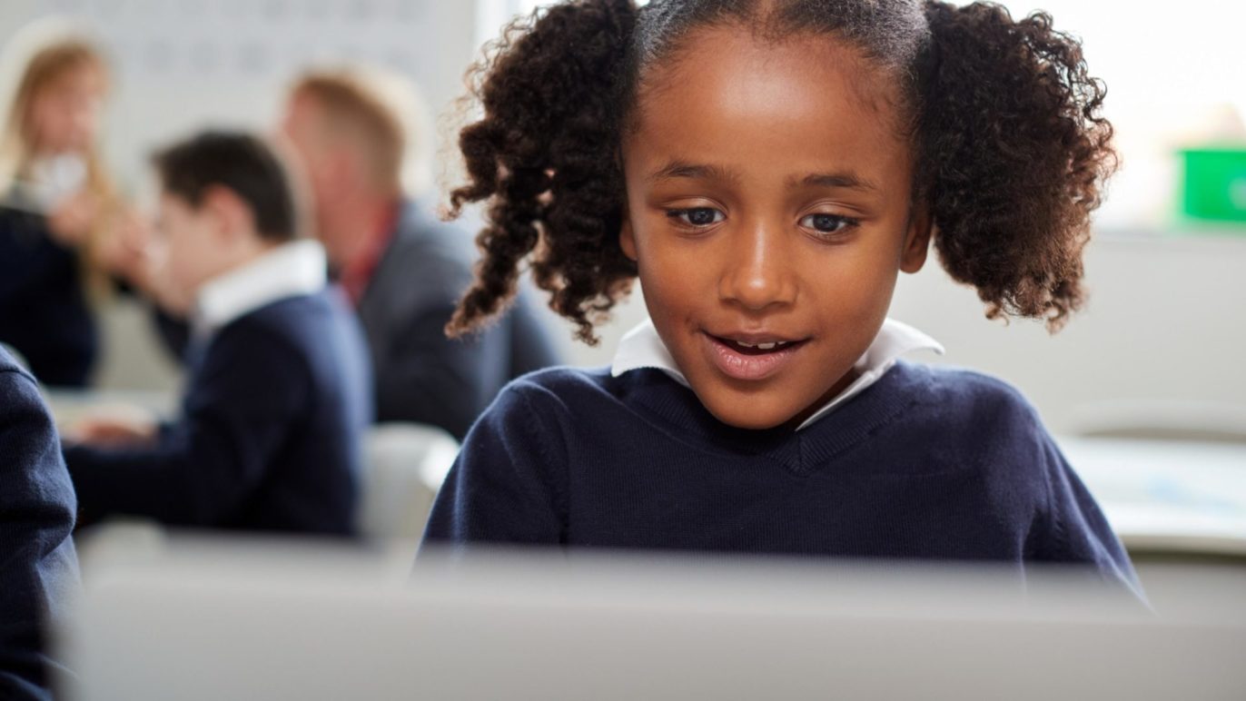 A photo of a black girl with pigtails sitting at a desk in a classroom, talking into a voice-enabled computer.