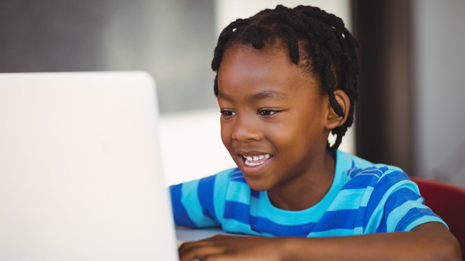 A photo of a young black boy wearing a striped blue t-shirt sitting at a desk, and talking into a voice-enabled laptop computer.