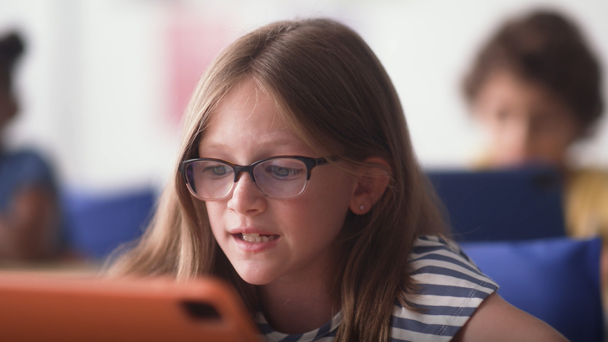 A photo of a young girl sitting at a desk in a classroom, talking into a computer.