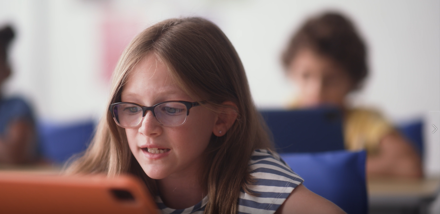 A photo of a girl sitting at a desk in a classroom with a tablet computer in front of her. She is practicing her reading fluency skills using a education app powered by voice AI.