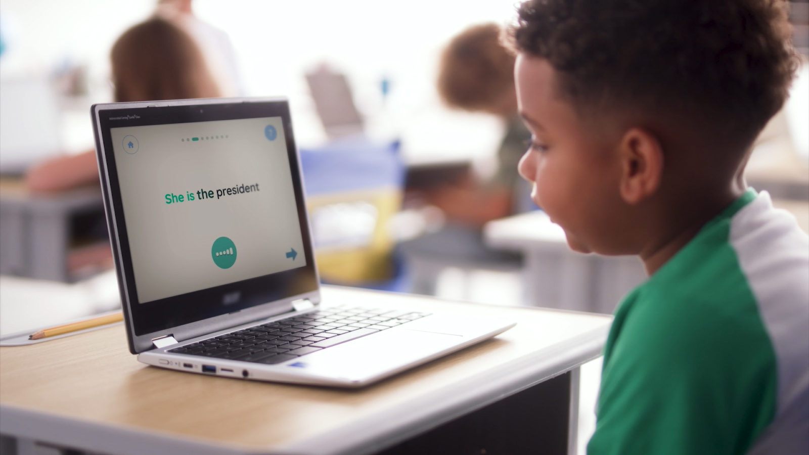 A photo of a boy sitting at a desk with a laptop in a classroom. He is practicing reading using a digital literacy app, powered by voice AI.