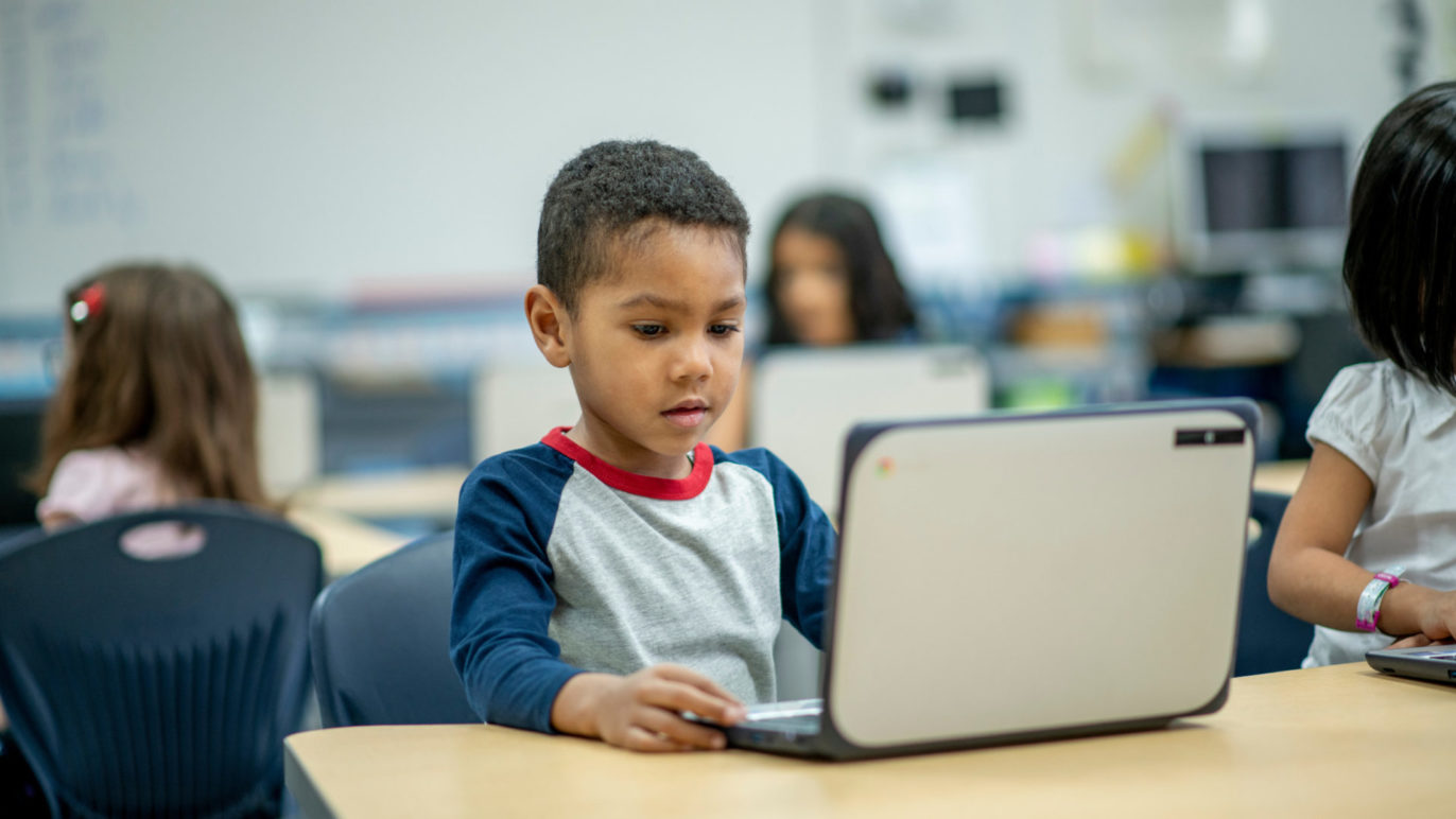 A photo of a young boy sitting at a desk in a classroom. He is completing a voice-activated reading activity on a laptop computer.