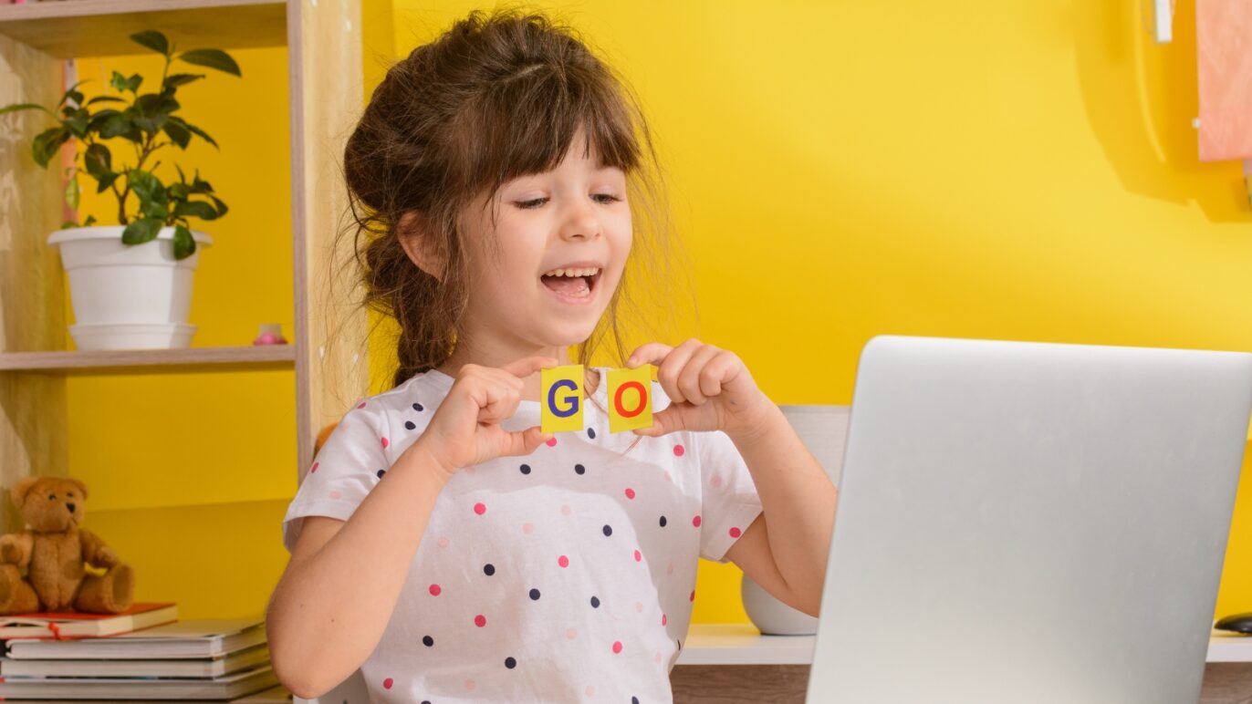 A photo of a girl sitting at a desk in front of a lap top computer. She is practicing her reading skills on a voice-enabled reading app.