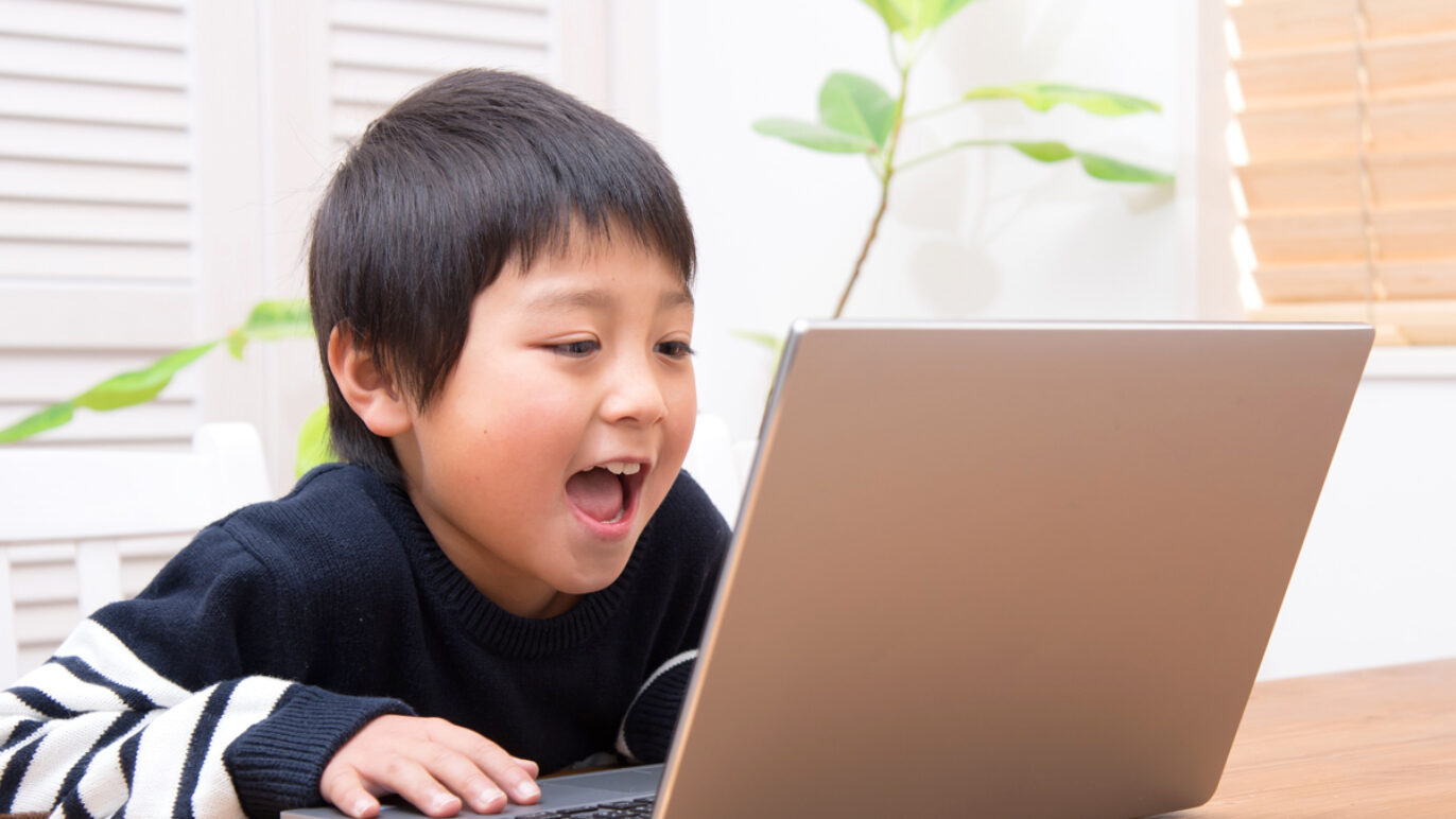 A photo of a young boy sitting at a desk speaking into his laptop computer.