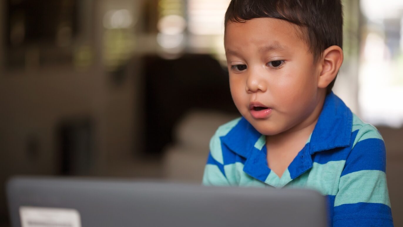 A photo of a young boy sitting at a desk, talking into a laptop computer. He is completing a voice-enabled literacy assessment, powered by SoapBox Labs' voice AI technology for kids.