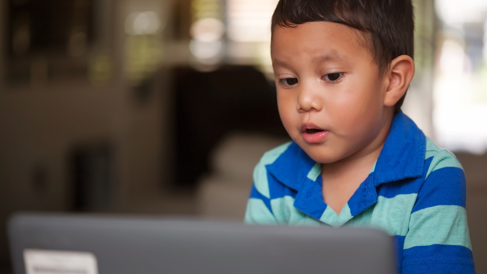 A photo of a young boy sitting at a desk, talking into a laptop computer. He is completing a voice-enabled literacy assessment, powered by SoapBox Labs' voice AI technology for kids.