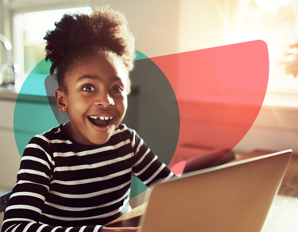 A photo of a girl sitting at a kitchen table on her laptop computer. She is smiling as she is completing a voice-enabled activity, powered by SoapBox Lab's equity-by-design speech recognition technology for kids.